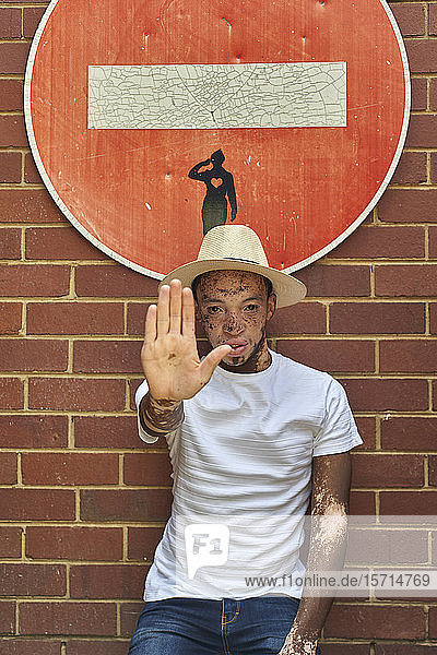 Porträt eines jungen Mannes mit Vitiligo  der einen Hut trägt und mit der Hand ein Stoppschild auf einem Verbotsschild macht