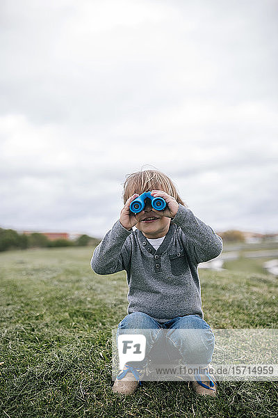 Smiling little boy crouching on a meadow looking through blue binoculars