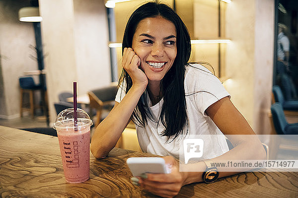 Schwarzhaarige Frau trinkt einen Smoothie und benutzt ein Smartphone im Café