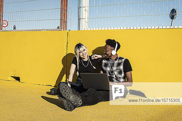 Lachendes junges Paar mit Kopfhörern und Laptop vor gelber Wand sitzend