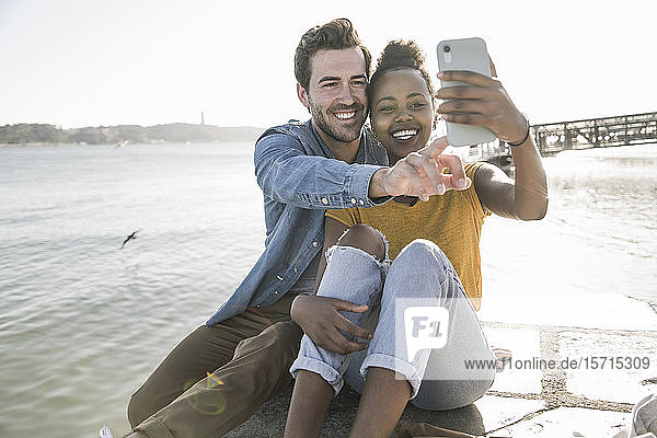 Happy young couple sitting on pier at the waterfront taking a selfie  Lisbon  Portugal