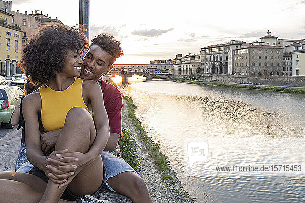 Liebenswertes junges Touristenpaar sitzt bei Sonnenuntergang auf einer Mauer am Arno  Florenz  Italien