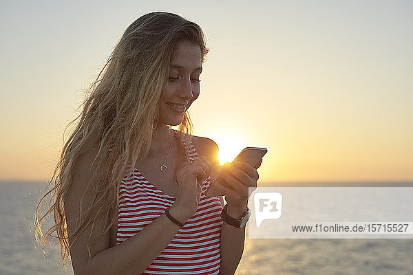 Junge Frau steht auf einer Klippe am Meer und benutzt ein Smartphone