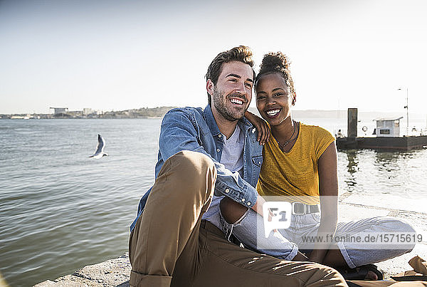 Happy young couple sitting on pier at the waterfront  Lisbon  Portugal