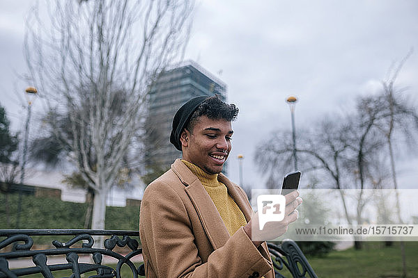 Lächelnder junger Mann sitzt auf einer Bank und benutzt sein Handy