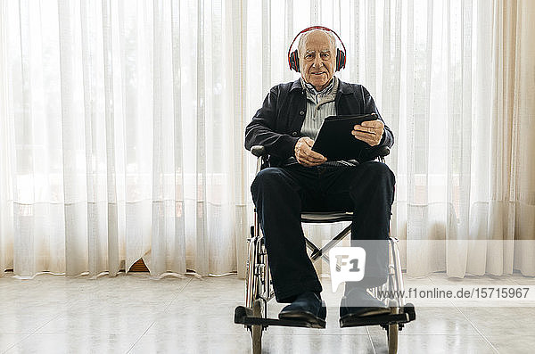 Porträt eines älteren Mannes im Rollstuhl sitzend mit digitalem Tablett  der mit Kopfhörern Musik hört