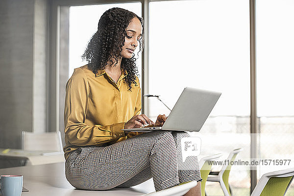 Young businesswoman sitting on desk in office using laptop