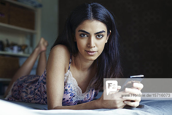 Porträt einer auf dem Bett liegenden jungen Frau mit Smartphone