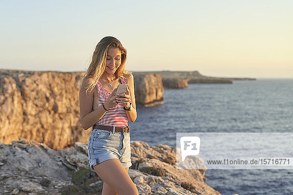 Junge Frau steht auf einer Klippe am Meer und benutzt ein Smartphone