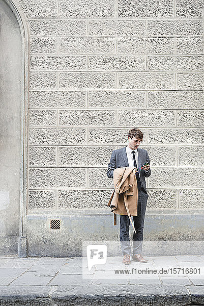 Junger Geschäftsmann steht auf Bürgersteig und schaut auf Smartphone