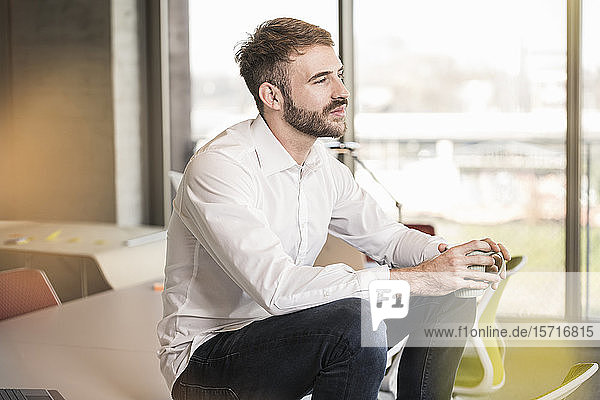 Young businessman sitting on conference table in office holding cup of coffee