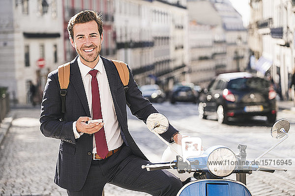Portrait of happy young businessman with cell phone and motor scooter in the city  Lisbon  Portugal