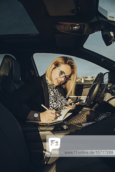 Junge blonde Frau  die im Auto ein Smartphone benutzt und in ein Notizbuch schreibt