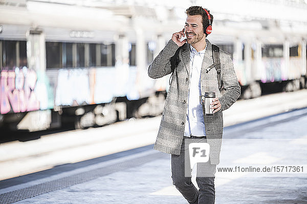 Glücklicher junger Geschäftsmann mit Kaffeetasse und Kopfhörern geht am Bahnhof spazieren