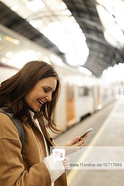 Glückliche junge Frau benutzt Mobiltelefon am Bahnhof