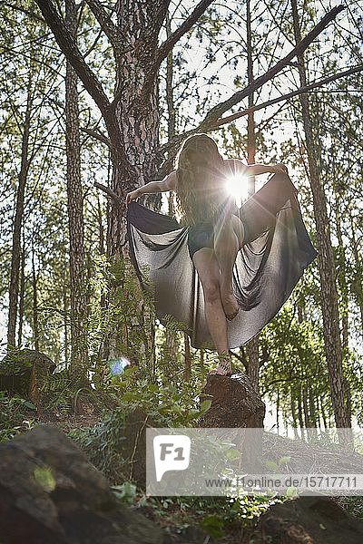 Barfüßige junge Frau steht auf einem Bein auf einem Felsen im Wald im Gegenlicht
