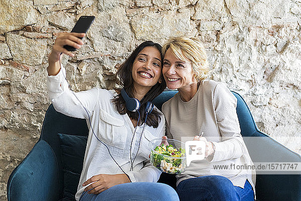Zwei glückliche Kollegen sitzen zur Mittagszeit auf der Couch und nehmen sich mit dem Smartphone ein