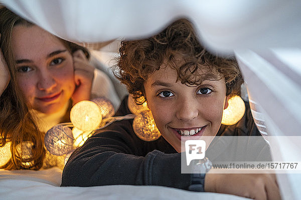 Porträt des glücklichen Geschwisterpaares mit Lichterkette unter der Bettdecke