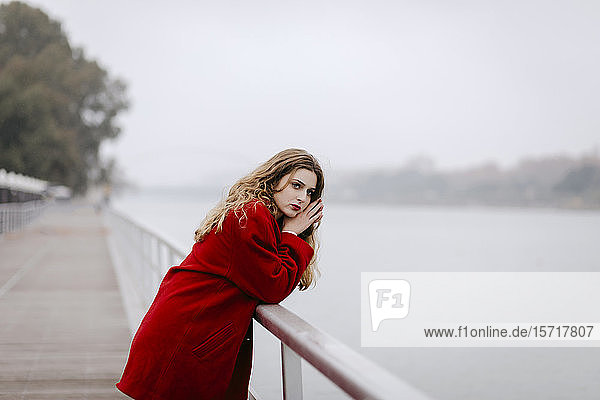 Porträt einer jungen Frau in rotem Mantel  die sich an einem regnerischen Tag an die Reling lehnt