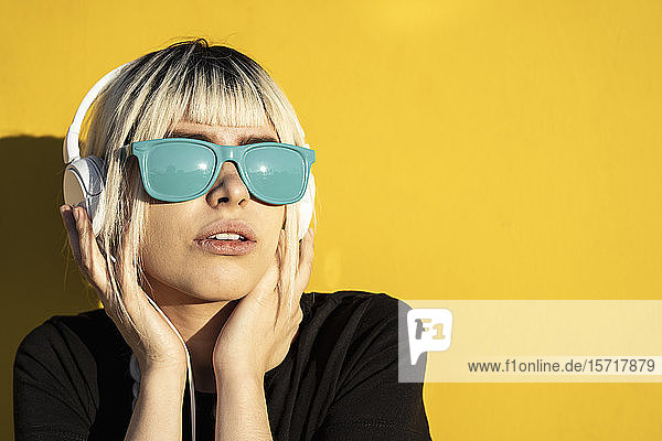 Porträt einer jungen Frau mit blauer Sonnenbrille  die vor einer gelben Wand Musik hört