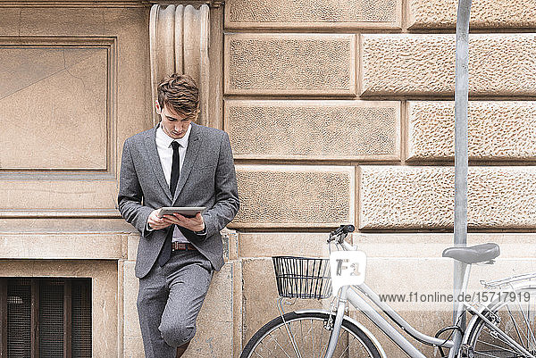 Junger Geschäftsmann lehnt sich an Fassade und schaut auf digitales Tablet