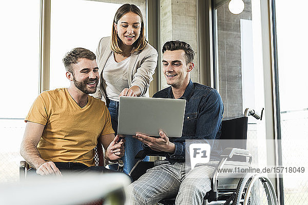 Junger Geschäftsmann im Rollstuhl zeigt Kollegen im Büro seinen Laptop