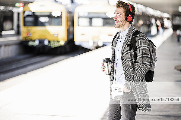 Lächelnder junger Geschäftsmann mit Kaffeetasse und Kopfhörer am Bahnhof