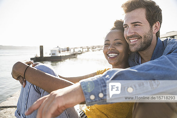 Portrait of happy young couple sitting on pier at the waterfront  Lisbon  Portugal