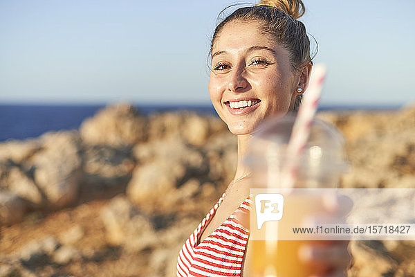 Junge Frau trinkt Saft am felsigen Strand