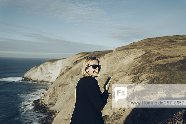 Rückansicht einer jungen blonden Frau mit Smartphone an der Küste