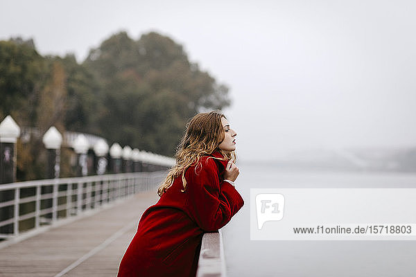Porträt einer jungen Frau in rotem Mantel  die sich an einem regnerischen Tag an die Reling lehnt