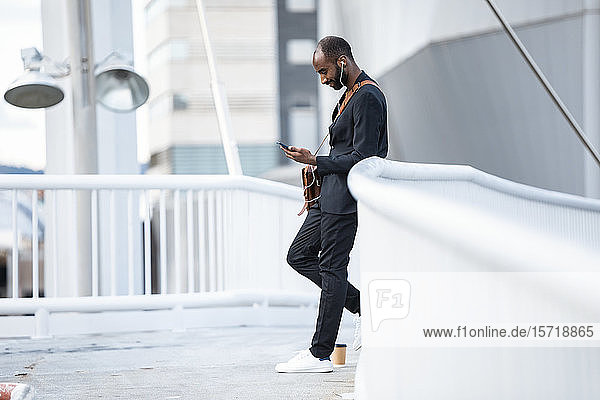 Junger Geschäftsmann mit Kopfhörern steht auf Steg und schaut auf Smartphone