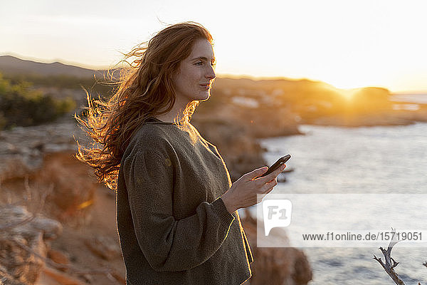 Rothaarige junge Frau mit Handy an der Küste bei Sonnenuntergang  Ibiza  Spanien
