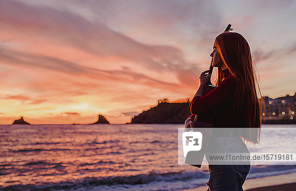 Junge Frau mit Gitarre  die bei Sonnenuntergang am Strand steht und auf das Meer schaut  Almunecar  Spanien