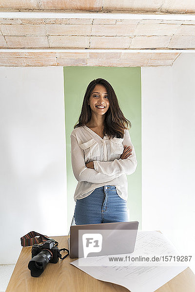 Portrait of smiling young architect with camera  laptop and construction plan on desk in a studio