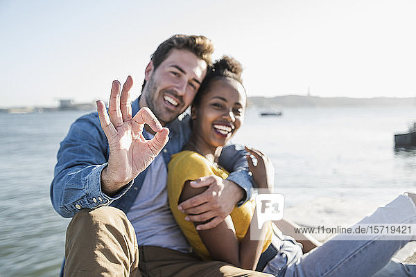 Portrait of happy young couple sitting at the waterfront gesturing  Lisbon  Portugal