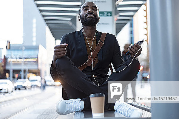Porträt eines jungen Geschäftsmannes mit Kopfhörern und Smartphone  der auf einer Bank an der Straßenbahnhaltestelle meditiert