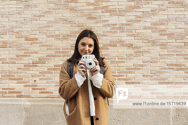 Porträt einer lächelnden jungen Frau mit Kamera vor einer Ziegelmauer
