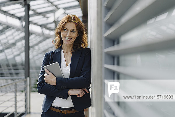 Porträt einer lächelnden Geschäftsfrau  die in einem modernen Bürogebäude ein Tablett hält