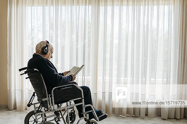 Senior man sitting in wheelchair reading a book while listening music with headphones