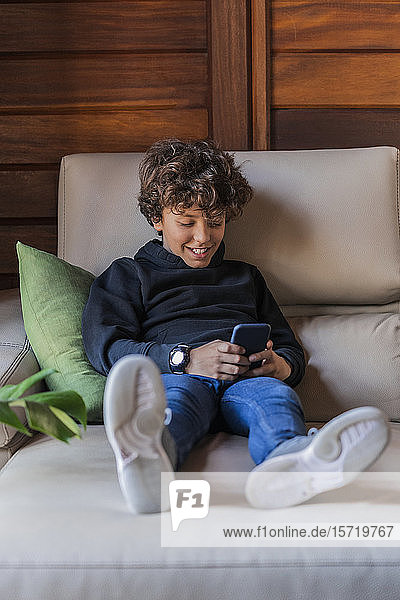 Lächelnder entspannter Junge  der sein Handy zu Hause auf der Couch benutzt