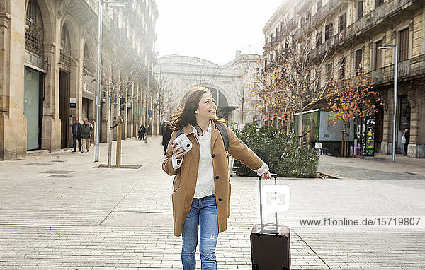 Lächelnde junge Frau mit Koffer und Kamera in der Stadt unterwegs  Barcelona  Spanien