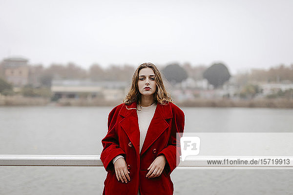 Porträt einer jungen Frau in rotem Mantel  die sich an einem Regentag an die Reling lehnt