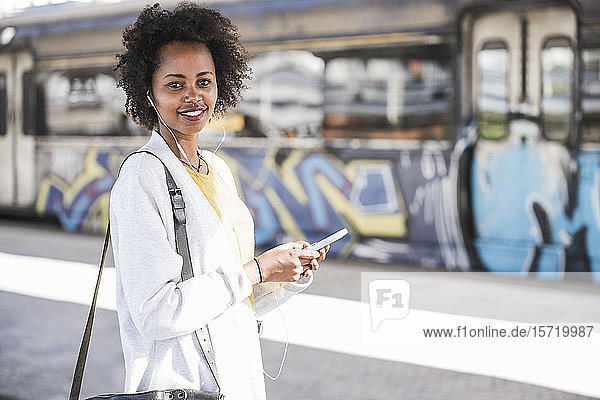 Porträt einer lächelnden jungen Frau mit Handy und Kopfhörer am Bahnhof