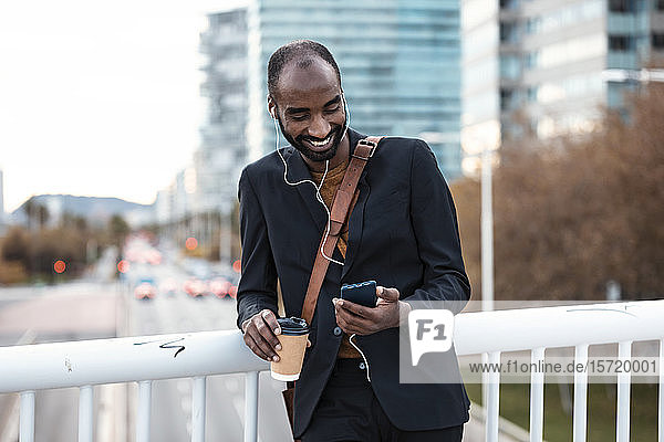 Lächelnder junger Geschäftsmann mit Kaffee zum Musikhören mit Kopfhörern und Smartphone
