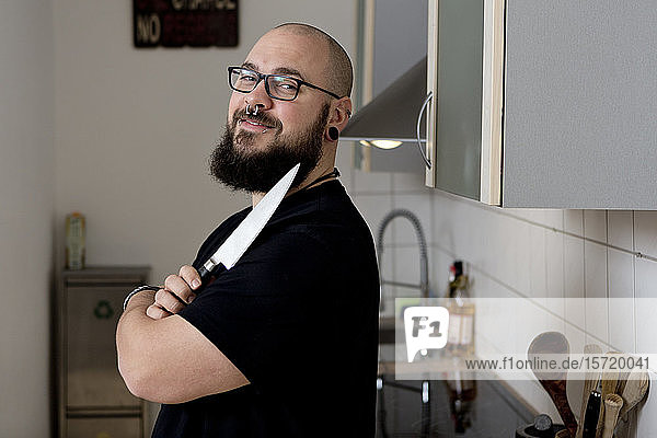 Porträt eines lächelnden bärtigen Mannes  der ein Küchenmesser in der Küche hält