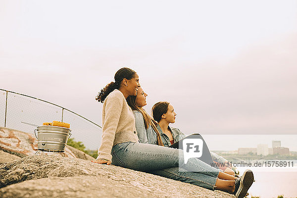 Glückliche Freundinnen schauen weg  während sie auf einer Felsformation am Seeufer gegen den Himmel sitzen