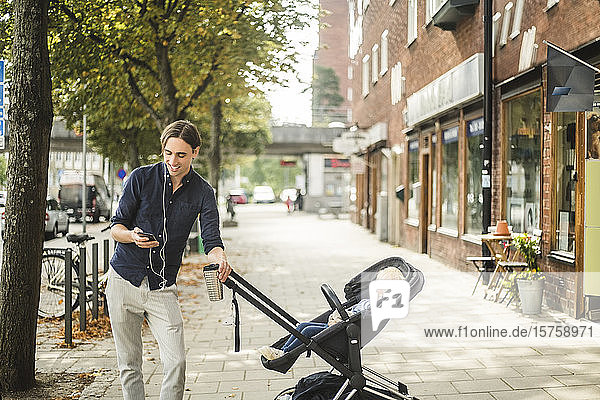 Smiling man using smart phone while standing by son on baby stroller in city