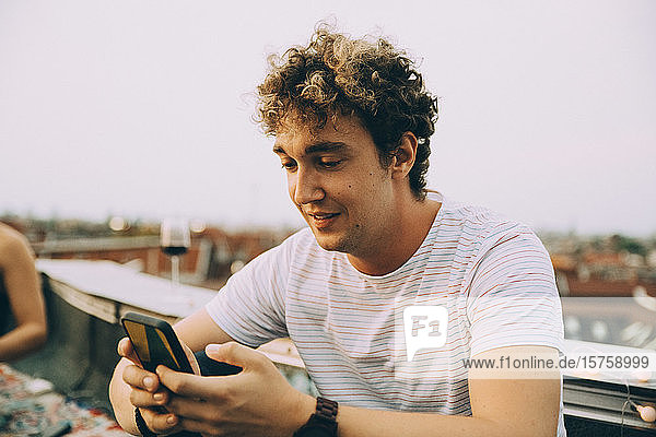 Junger Mann verschickt Textnachrichten per Smartphone  während er auf einer Terrasse in der Stadt sitzt