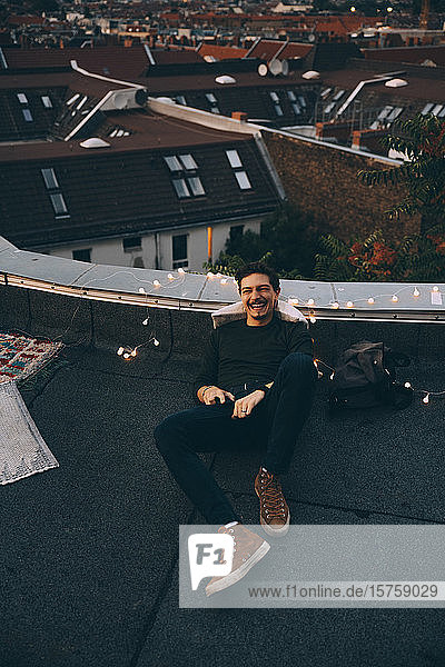 Hochwinkel in voller Länge eines glücklichen jungen Mannes der sich während einer Party in der Abenddämmerung auf der beleuchteten Terrasse entspannt